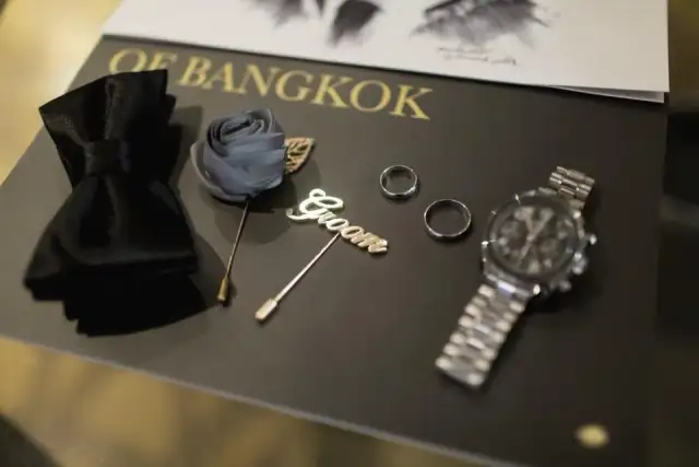 A black bow tie, a lapel pin, two rings and a steel watch arranged on a dark presentation box