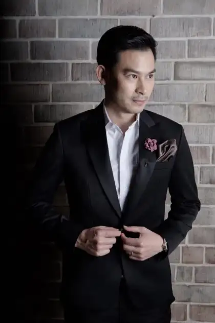 A man in a black jacket with satin lapels, a flower lapel pin and a silk pocket square, brick behind