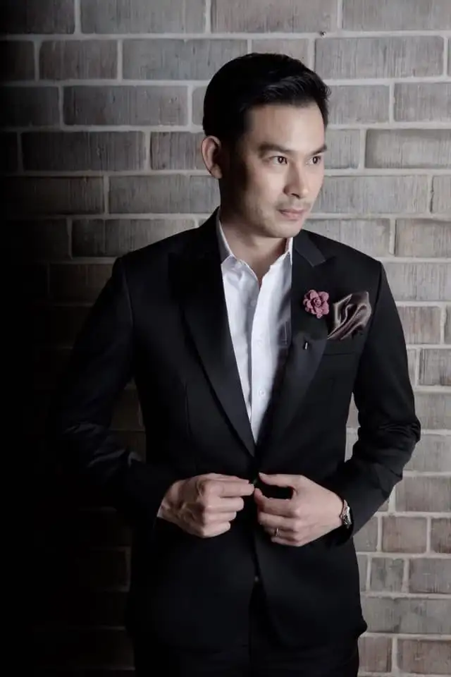 A man in a black jacket with satin lapels, a flower lapel pin and a silk pocket square, brick behind
