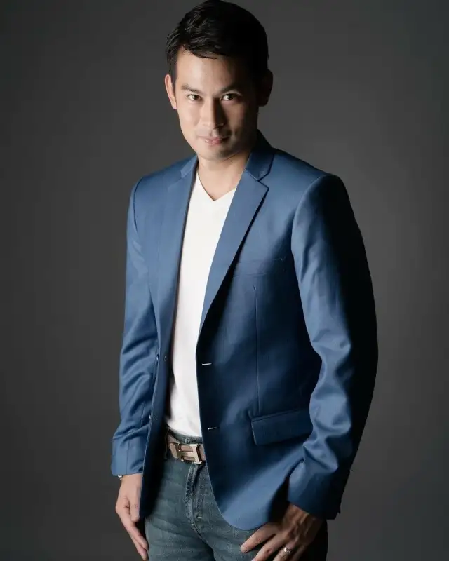 A man in a blue blazer over a white T-shirt and jeans against a dark grey studio backdrop