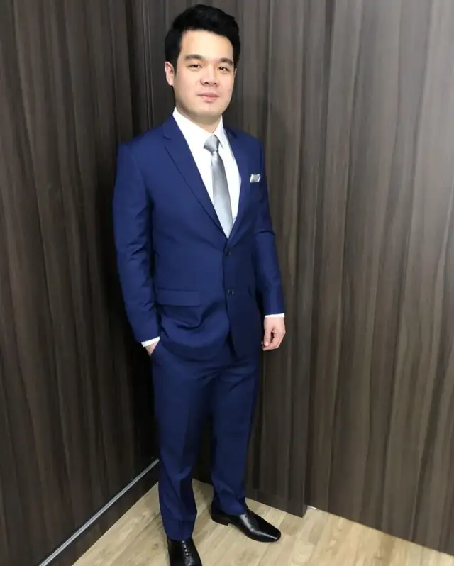 A man in a blue suit standing full length against a dark wall