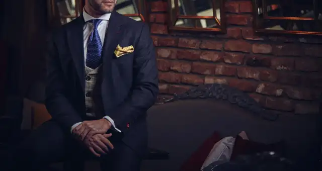 A man in a dark suit and gold bow tie against a brick wall