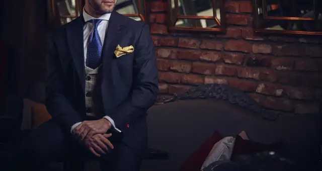 A man in a dark suit and gold bow tie against a brick wall