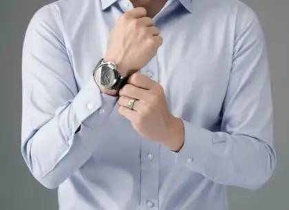 A man in a pale blue shirt adjusting the watch at his cuff, shirt sleeve and cuff button in focus