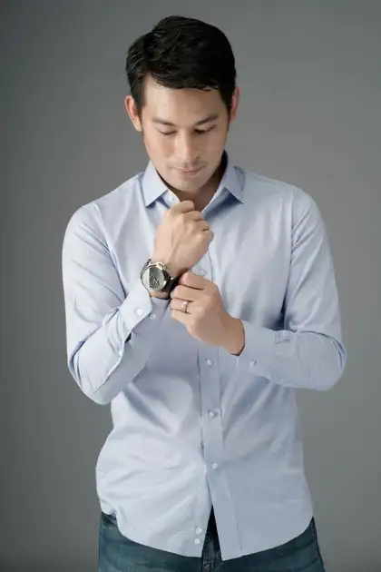 A man in a pale blue shirt fastening his cuff against a grey studio backdrop