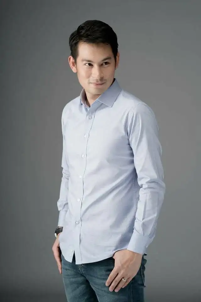 A man in a pale blue shirt and jeans, hand in his pocket, against a grey studio backdrop