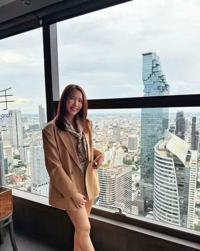 A woman in a tan jacket standing at a window above the city